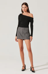A woman is wearing a black, off-the-shoulder, long-sleeve top with draped detailing on one shoulder. She pairs it with a high-waisted, gray plaid mini skirt. She completes the outfit with black high-heeled shoes with ankle straps.